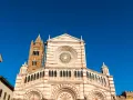 Il Duomo di Grosseto, con la sua facciata a strisce bianche e rosa e il rosone centrale, si staglia contro un cielo azzurro intenso.