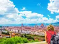 Vista mozzafiato di Firenze dal Piazzale Michelangelo. Una donna ammira il Duomo e il Ponte Vecchio sotto un cielo azzurro con nuvole bianche.
