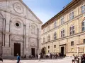 Piazza Pio II a Pienza, con la facciata del Duomo e il Palazzo Piccolomini. Turisti passeggiano nella piazza soleggiata.
