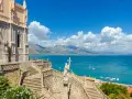 La chiesa domina il mare azzurro della Riviera di Ulisse. Scale imponenti conducono a un portale gotico, mentre barche solcano le acque.