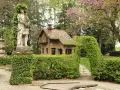 Giardino incantato a Grazzano Visconti con statua romana, casetta in mattoni e sentiero tra siepi fiorite.
