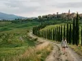 Una persona cammina lungo la **Val d'Orcia route**, circondata da dolci colline verdi e cipressi, con un borgo medievale sullo sfondo.