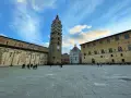 Piazza del Duomo di Pistoia con la torre campanaria, il battistero e il palazzo vescovile sotto un cielo nuvoloso.