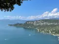 Vista panoramica del Lago di Garda con San Zeno di Montagna sullo sfondo, tra montagne verdi e cielo azzurro.