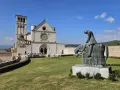 Statua di un cavaliere pensieroso davanti alla maestosa basilica San Francesco d'Assisi, sotto un cielo azzurro.