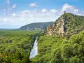 Un fiume serpeggia tra la fitta vegetazione del Parco dell'Uccellina, con una torre antica che domina la collina rocciosa.