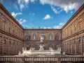 Cortile del Palazzo Pitti con fontana e statue, sotto un cielo azzurro con nuvole bianche.
