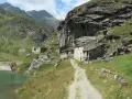 Antico borgo in pietra nel **Val di Susa**, con un sentiero che sale verso le case arroccate su un pendio verdeggiante, vicino a un lago alpino.