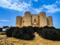 Castel del Monte, un'icona pugliese, si erge maestoso su una collina, circondato da una vegetazione rigogliosa sotto un cielo azzurro.