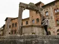 Donna sorride vicino all'arco in pietra e al pozzo di San Gimignano, con palazzi medievali sullo sfondo.