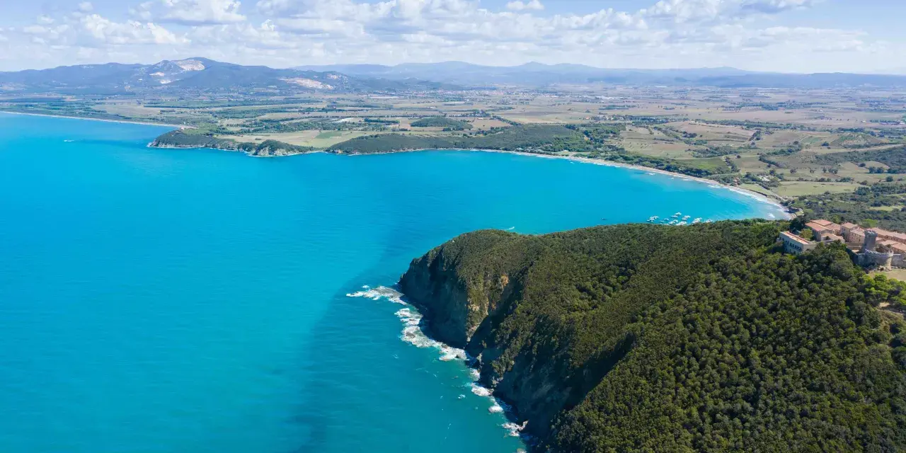 Vista aerea della costa degli Etruschi con mare azzurro, scogliere verdi e un borgo storico.