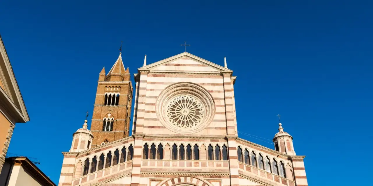 Il Duomo di Grosseto, con la sua facciata a strisce bianche e rosa e il rosone centrale, si staglia contro un cielo azzurro intenso.