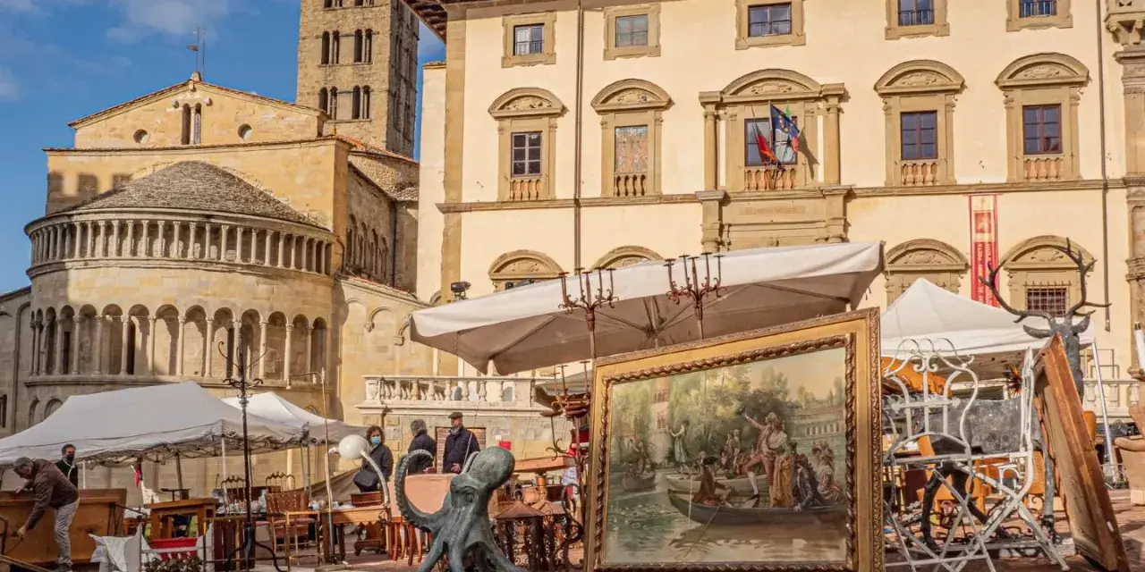 Mercatino d'antiquariato in piazza Grande ad Arezzo, con opere d'arte, sculture e mobili esposti sotto il sole.