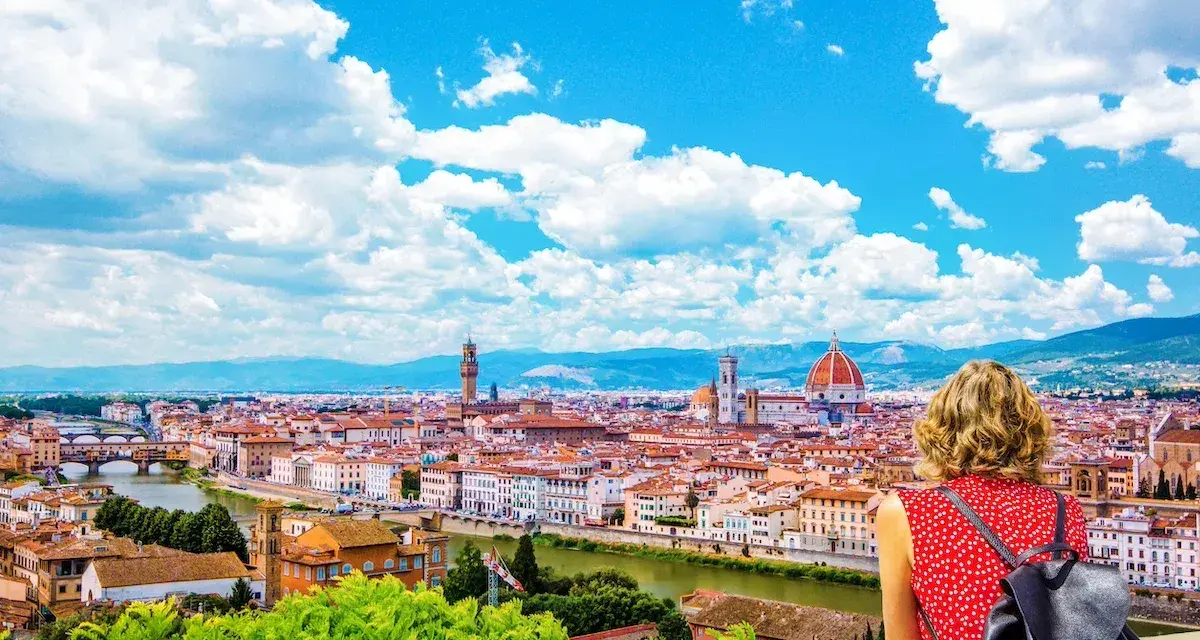 Vista mozzafiato di Firenze dal Piazzale Michelangelo. Una donna ammira il Duomo e il Ponte Vecchio sotto un cielo azzurro con nuvole bianche.