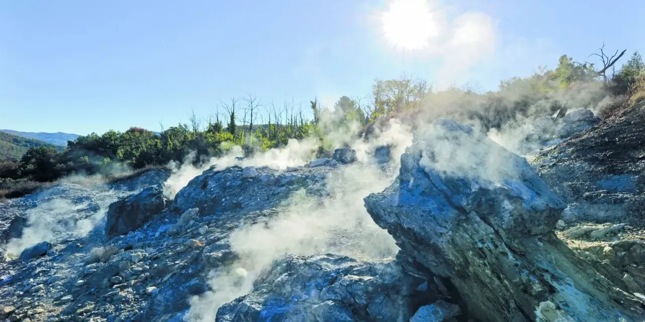 Fumarole attive nella valle del diavolo, con rocce grigie e blu avvolte dal vapore. Il sole splende nel cielo azzurro.