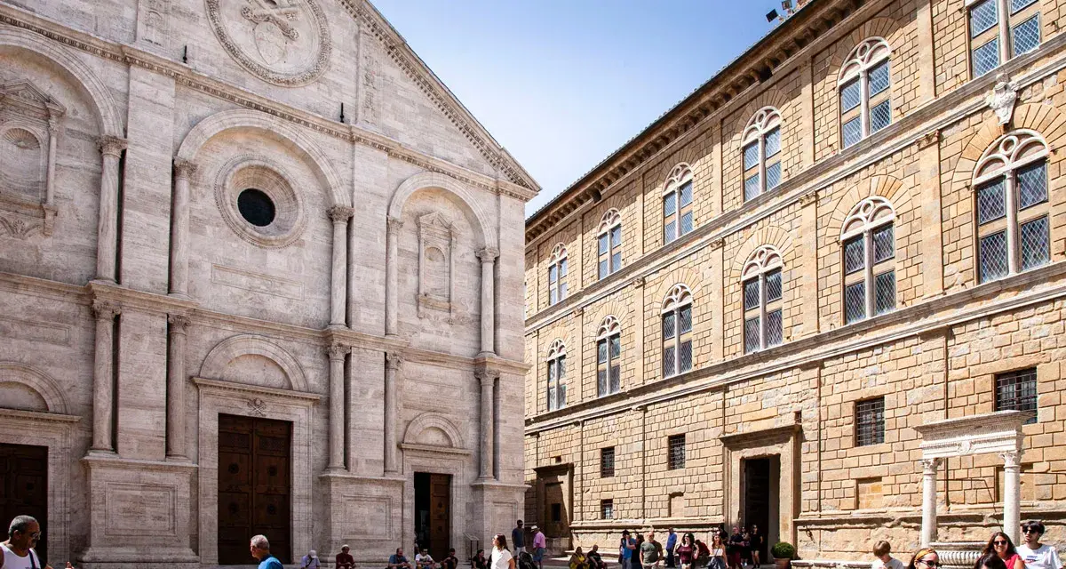 Piazza Pio II a Pienza, con la facciata del Duomo e il Palazzo Piccolomini. Turisti passeggiano nella piazza soleggiata.