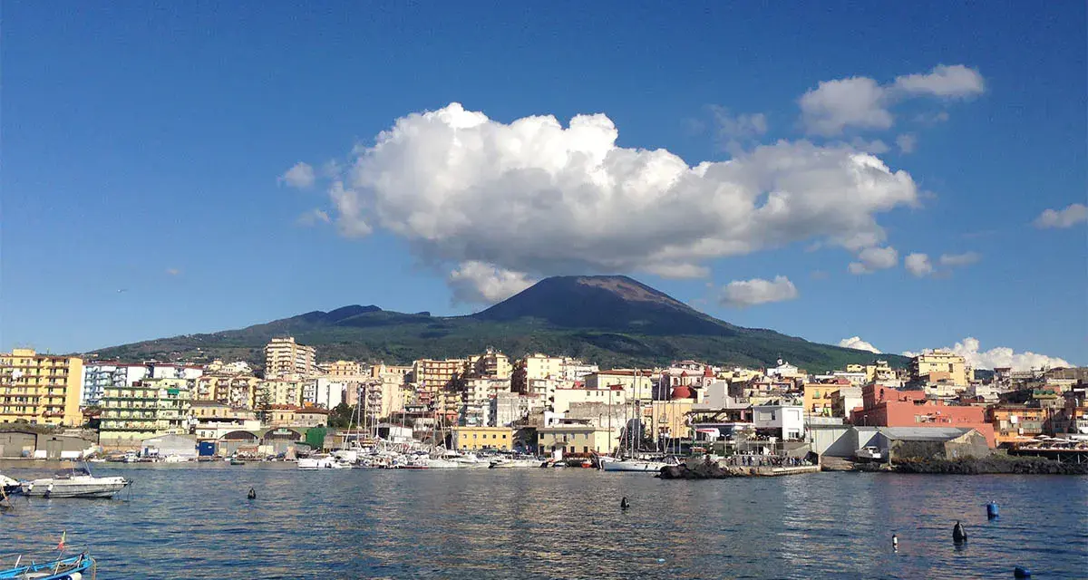 Vista del porto di Torre del Greco con il Vesuvio sullo sfondo, sotto un cielo azzurro con nuvole bianche.