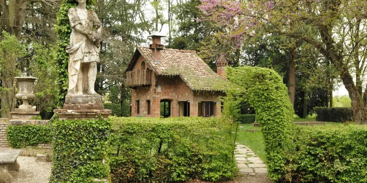 Giardino incantato a Grazzano Visconti con statua romana, casetta in mattoni e sentiero tra siepi fiorite.