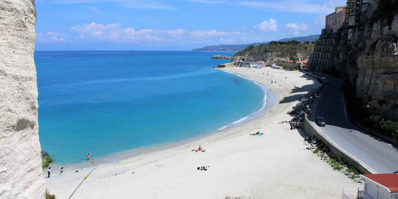 Spiaggia bianca e mare cristallino sulla **Costa degli Dei**. Case aggrappate alla scogliera, una strada panoramica e bagnanti che si godono il sole.