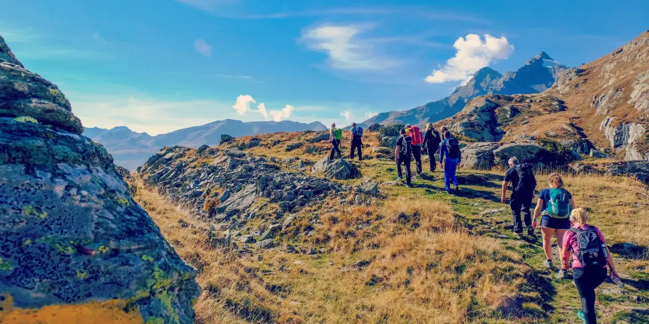 Un gruppo di escursionisti cammina su un sentiero roccioso e erboso, con montagne sullo sfondo. Il **Passo dello Spluga** offre panorami mozzafiato.