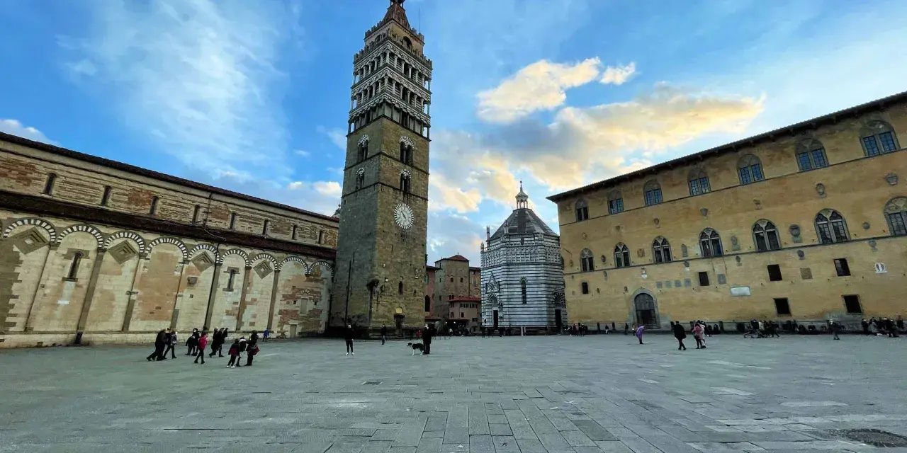 Piazza del Duomo di Pistoia con la torre campanaria, il battistero e il palazzo vescovile sotto un cielo nuvoloso.