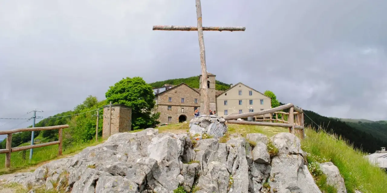 Croce di legno a San Pellegrino in Alpe, con edifici storici sullo sfondo e rocce in primo piano.