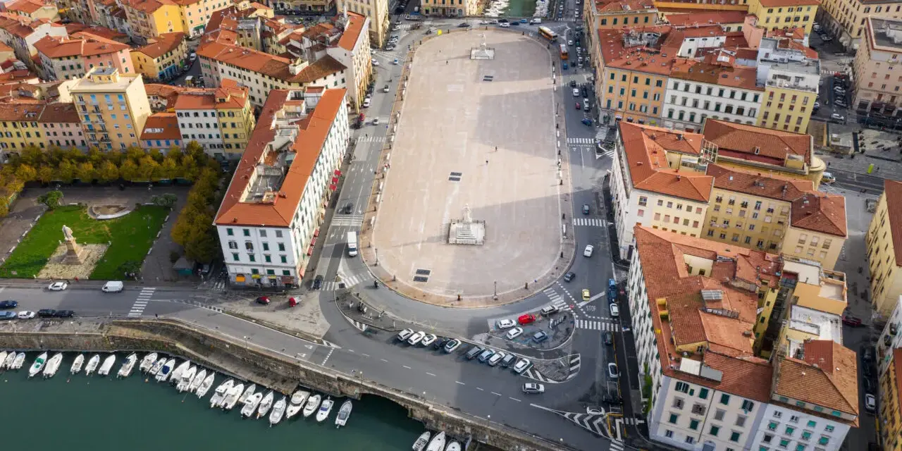 Vista aerea della **Piazza della Repubblica a Livorno**, con edifici storici, un monumento centrale, auto e barche ormeggiate nel canale.