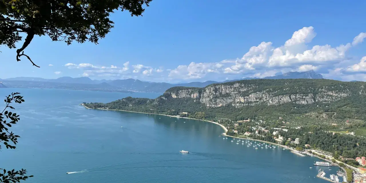 Vista panoramica del Lago di Garda con San Zeno di Montagna sullo sfondo, tra montagne verdi e cielo azzurro.