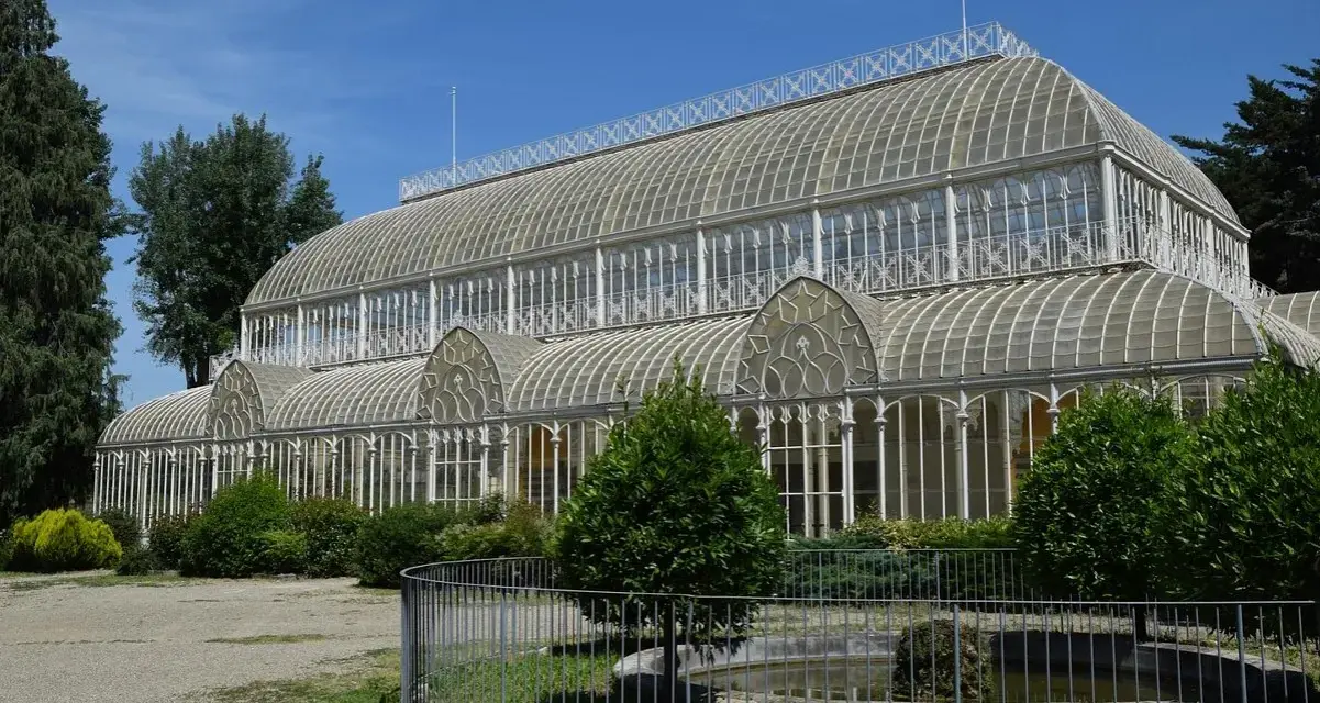 Serra in vetro con archi decorati, parte del giardino orticoltura firenze, circondata da alberi e vegetazione.
