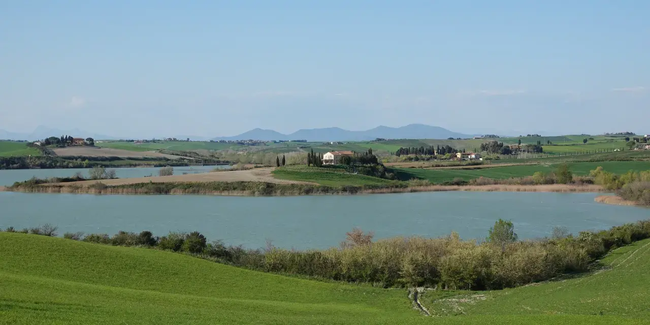 Campagna toscana con lago e casale in collina, un luogo dove la **santa luce cosa vedere** è ovunque, tra dolci pendii verdi e il cielo azzurro.