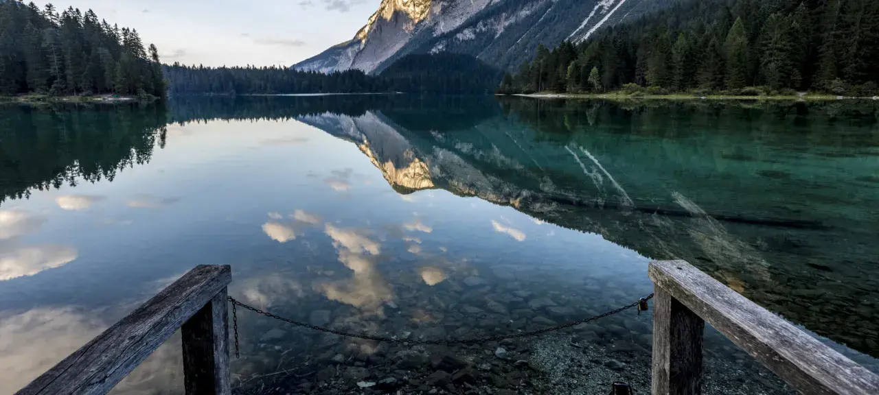 Lago alpino con riflessi di montagne e nuvole, un pontile in legno invita a contemplare la bellezza del Val di Non.