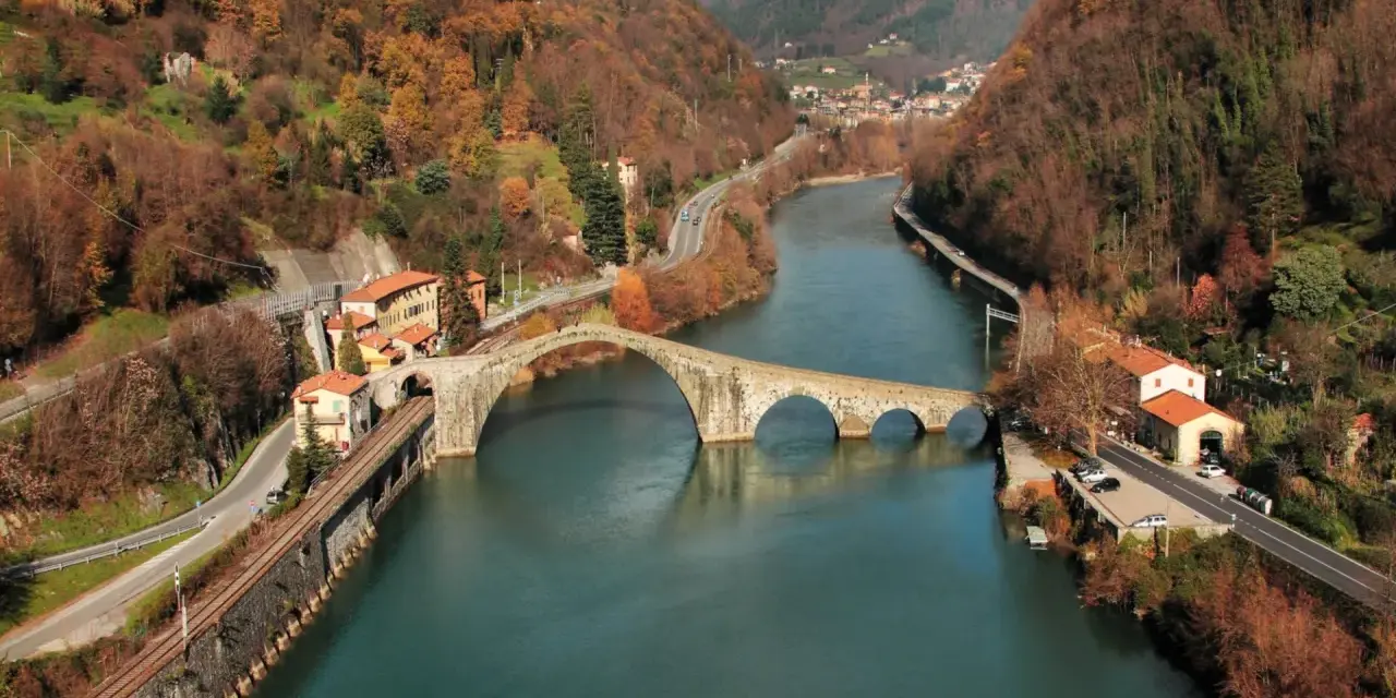 Il **ponte della Maddalena**, con i suoi archi irregolari, attraversa un fiume circondato da colline autunnali e case colorate.