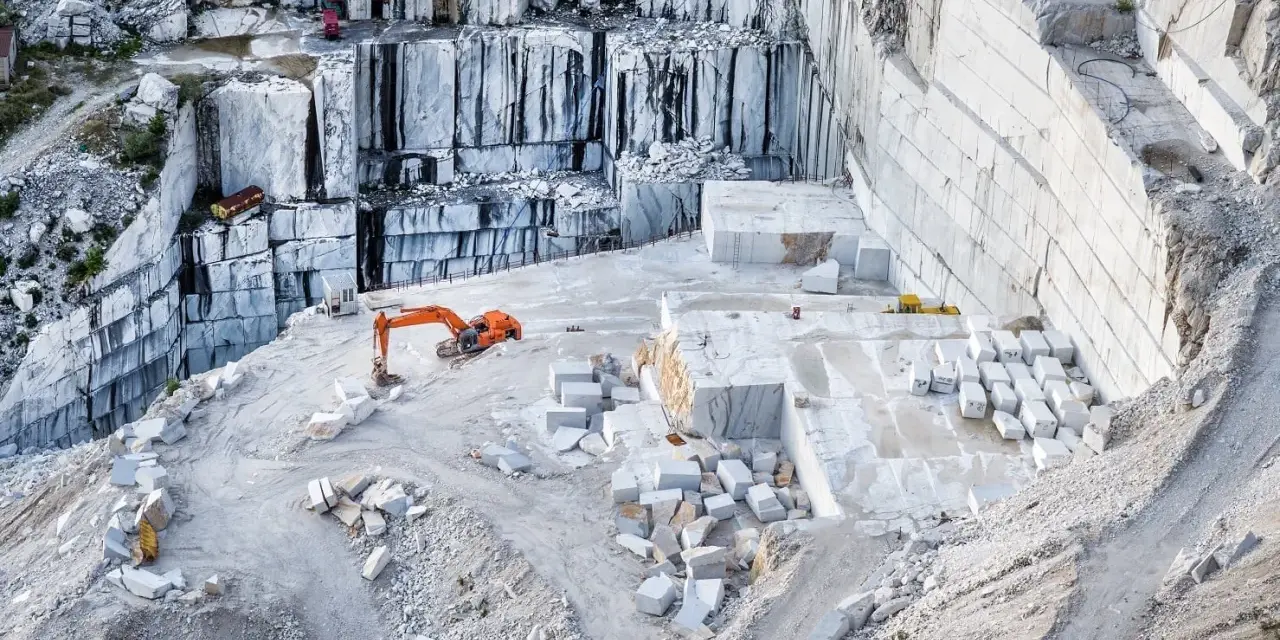 Cava di marmo a Carrara, con escavatore arancione al lavoro tra blocchi di pietra bianca. Un luogo affascinante da vedere a Carrara.