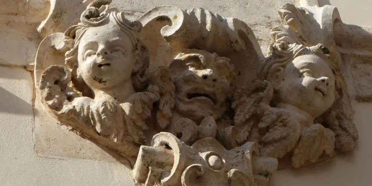 Dettaglio scultoreo barocco a Martina Franca: volti di putti e una maschera grottesca emergono dalla pietra calcarea, testimoni di un'antica arte.