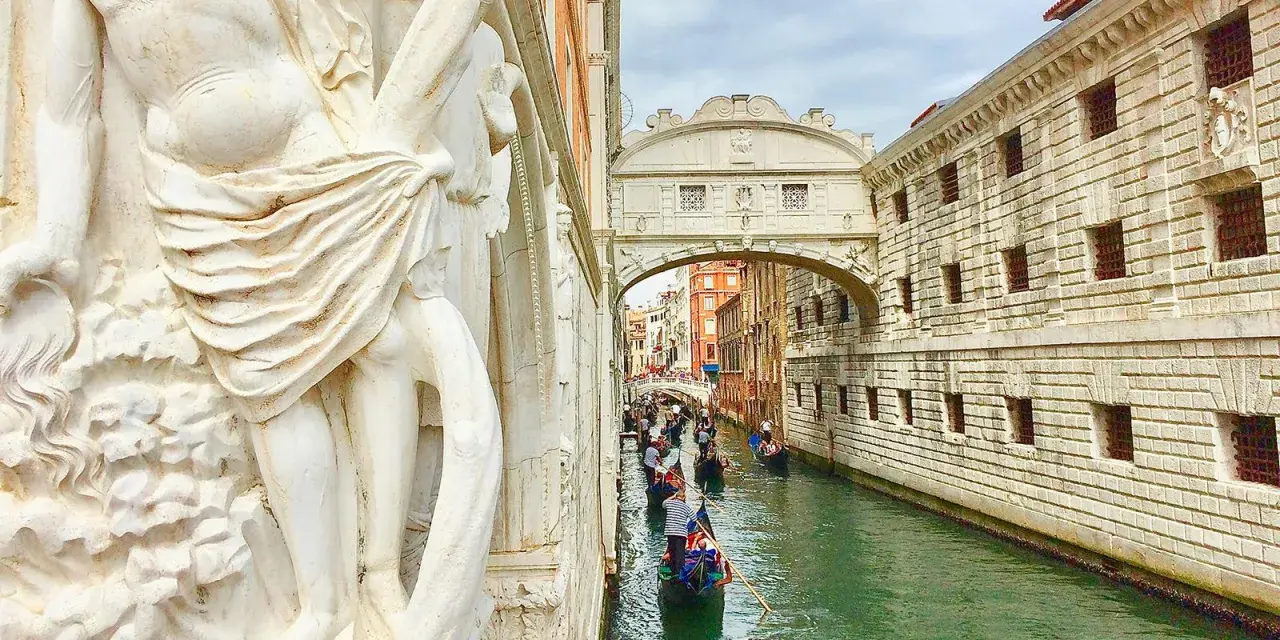Gondole scivolano sul canale sotto il Ponte dei Sospiri, con un'imponente statua in primo piano.