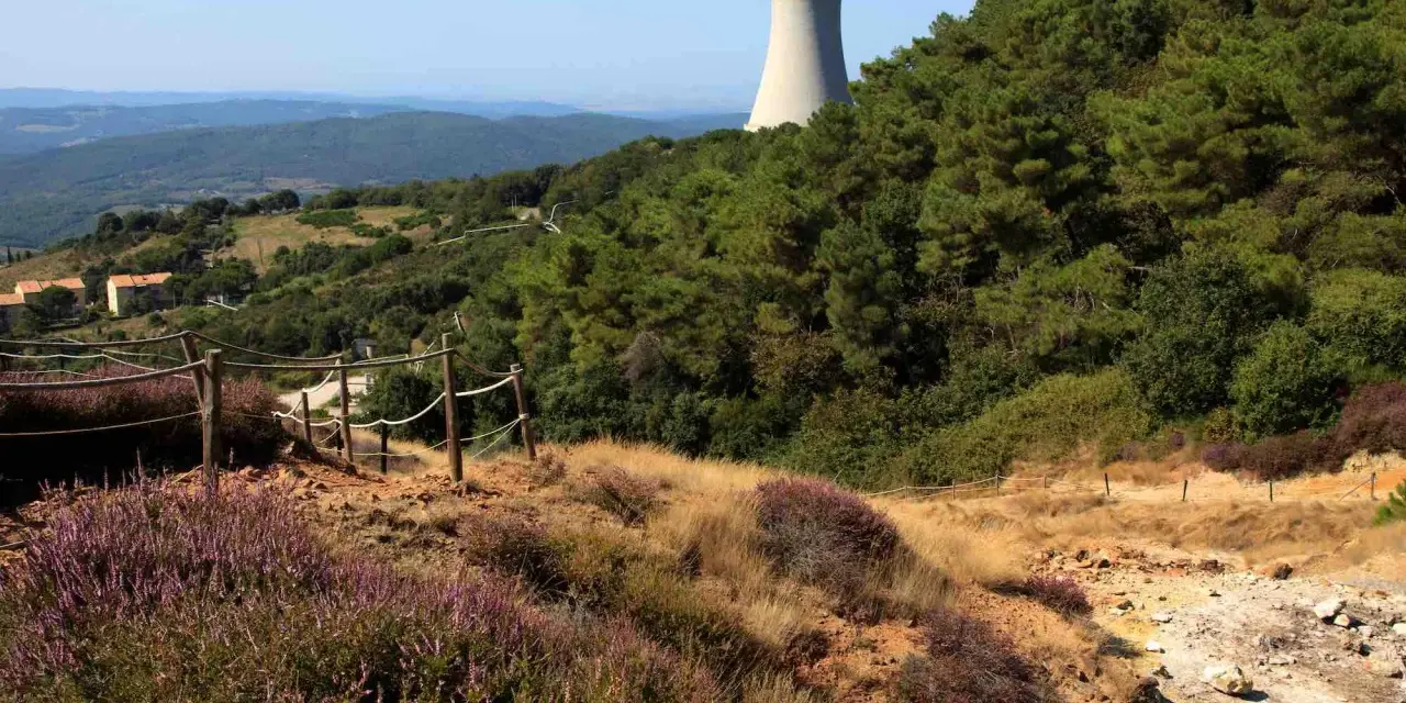 Sentiero nel parco delle Biancane con torretta di raffreddamento in lontananza, circondato da vegetazione e colline.