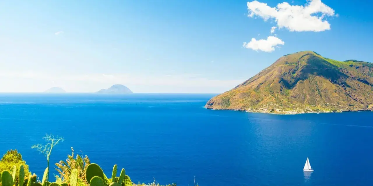 Panoramica delle Isole Eolie: cosa vedere? Un veliero solca il blu intenso del mare, con isole vulcaniche sullo sfondo e fichi d'India in primo piano.