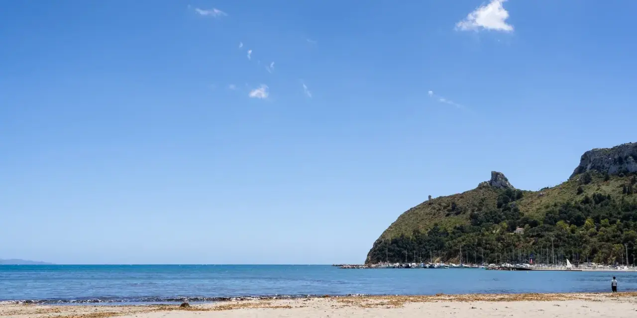 Spiaggia del Poetto con mare azzurro, montagne verdi e un porto turistico pieno di barche.