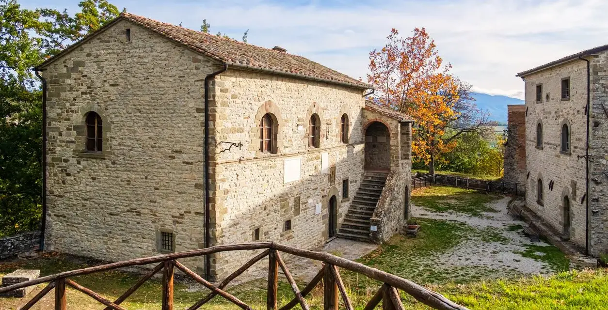 Antica strada in Casentino conduce a un borgo in pietra con un cortile interno e un albero dalle foglie arancioni.