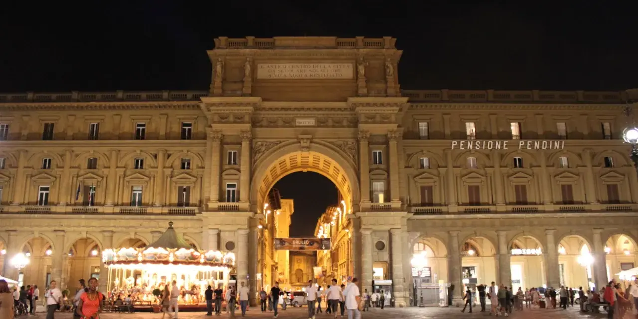 La piazza della Repubblica a Firenze di notte, con il suo arco monumentale illuminato, gente che passeggia e una giostra scintillante.