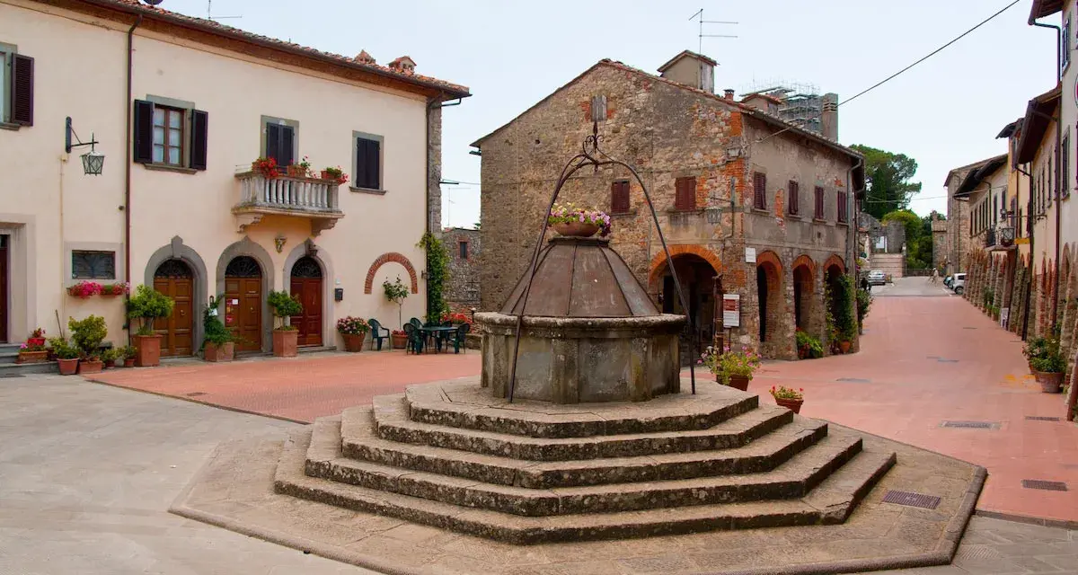 Piazza con pozzo antico a Civitella in Val di Chiana, circondata da case in pietra e intonaco, con fiori colorati.