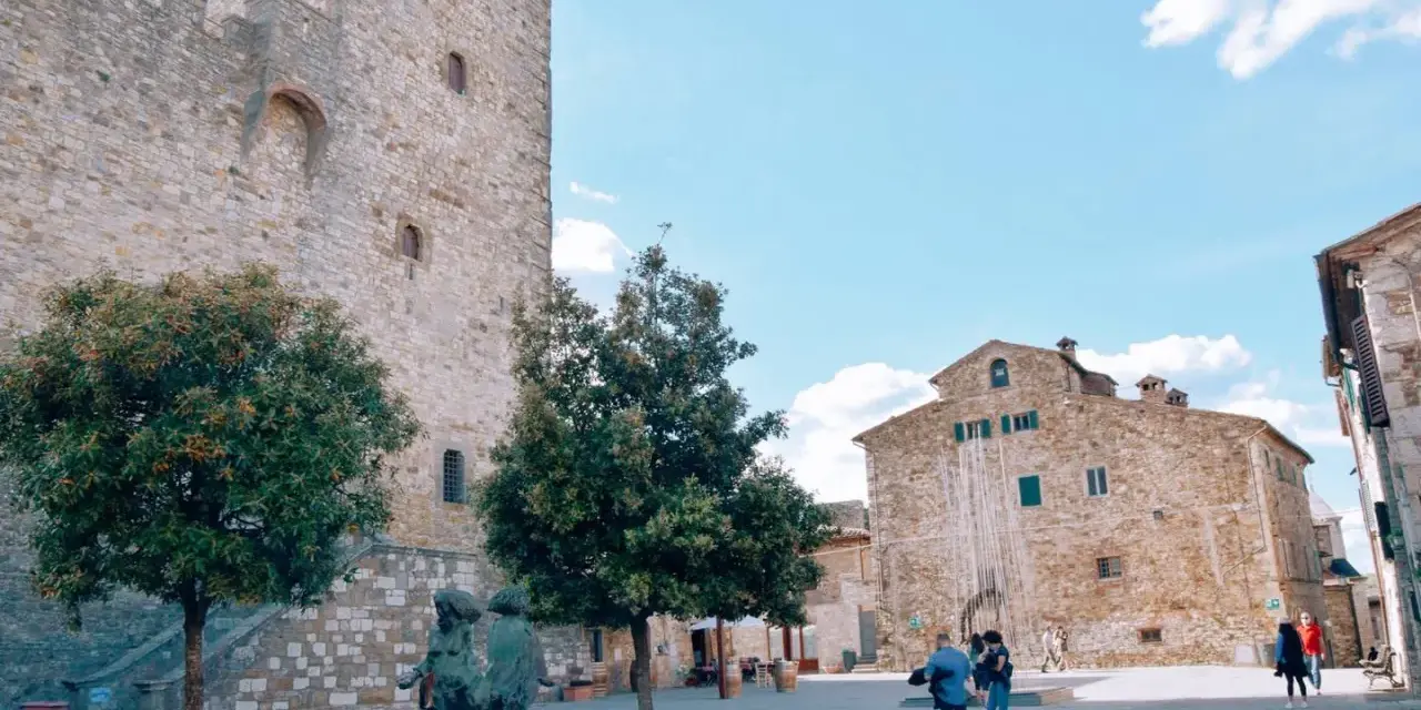 Piazza nel centro storico di Castellina in Chianti con torre medievale, edifici in pietra e scultura.
