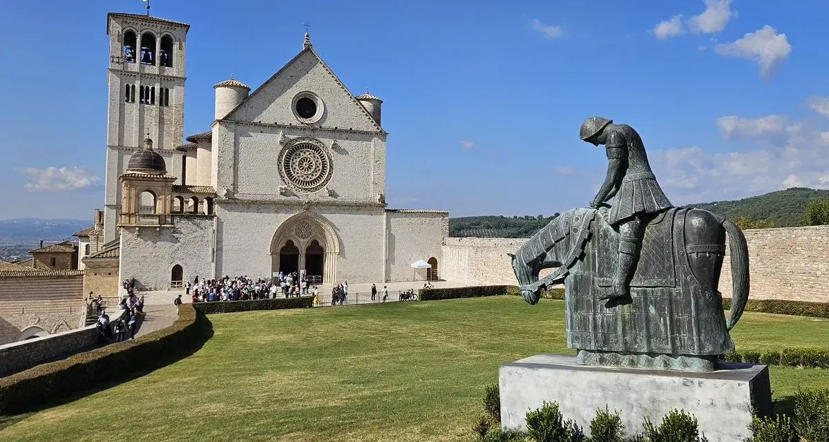 Statua di un cavaliere pensieroso davanti alla maestosa basilica San Francesco d'Assisi, sotto un cielo azzurro.