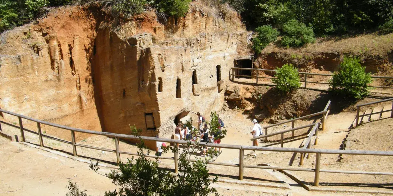 Antiche cave di tufo scavate nella roccia, con finestre e ingressi. Un gruppo di turisti esplora questo luogo affascinante, tra i **baratti cosa vedere** e la natura rigogliosa.
