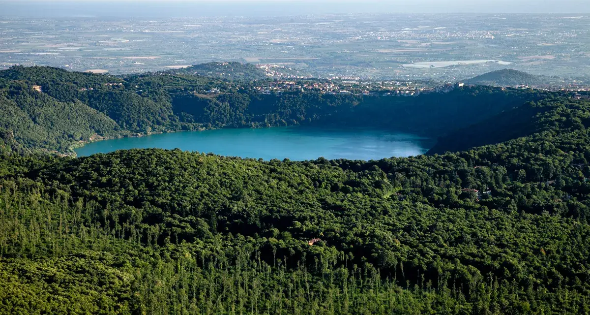 Vista panoramica del lago di Albano, circondato da rigogliosa vegetazione, con il borgo di Rocca di Papa in lontananza.