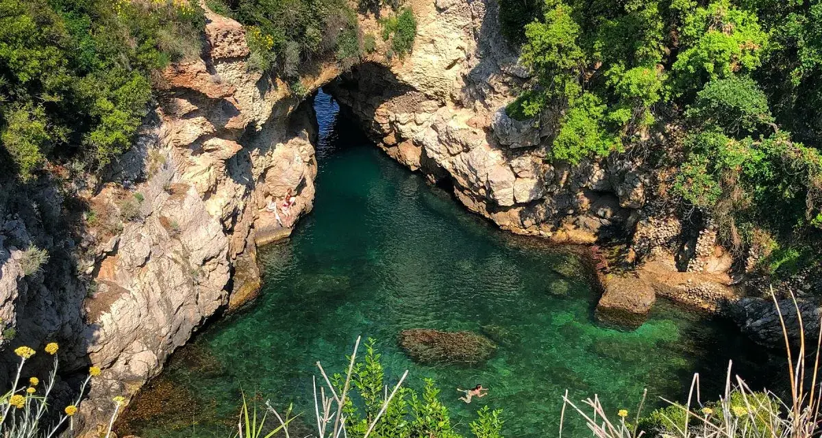 Bagni Regina Giovanna: una baia nascosta con acque cristalline, rocce scolpite e vegetazione lussureggiante. Persone si godono il sole e nuotano.