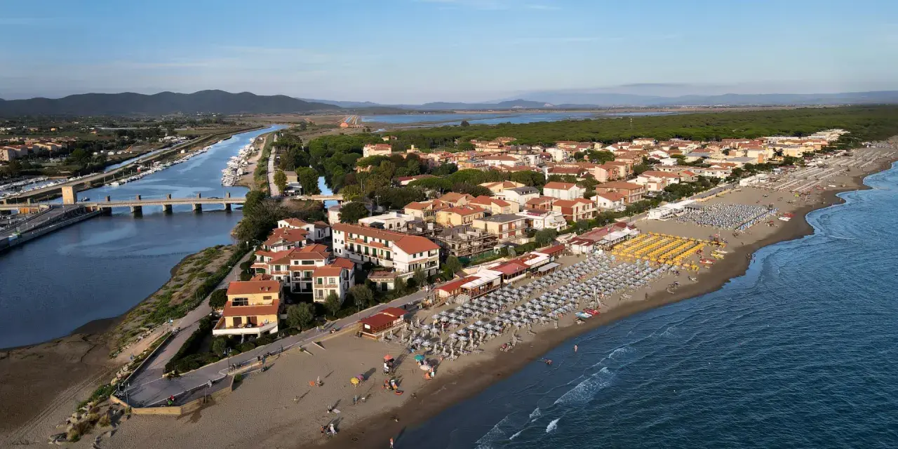 Vista aerea di Castiglione della Pescaia: un canale navigabile con barche, case con tetti rossi e una spiaggia attrezzata con ombrelloni e lettini, bagnata dal mare.