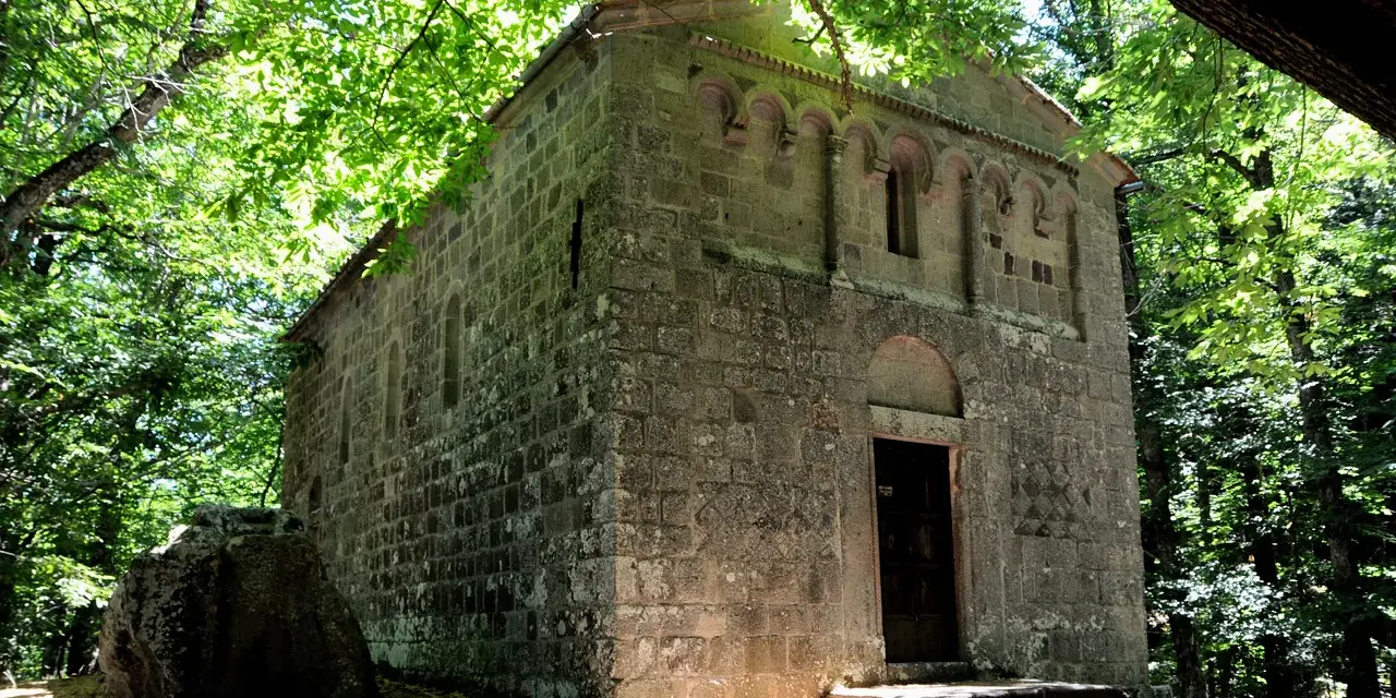 Antica pieve in pietra nel bosco Isabella, Radicofani. La luce filtra tra gli alberi illuminando la facciata.