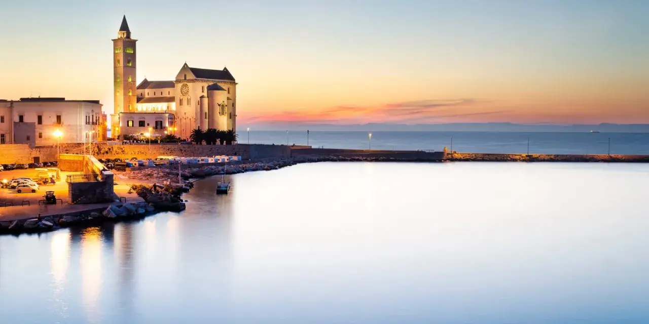 La cattedrale di Trani si erge maestosa sul mare al tramonto, illuminata da luci calde.