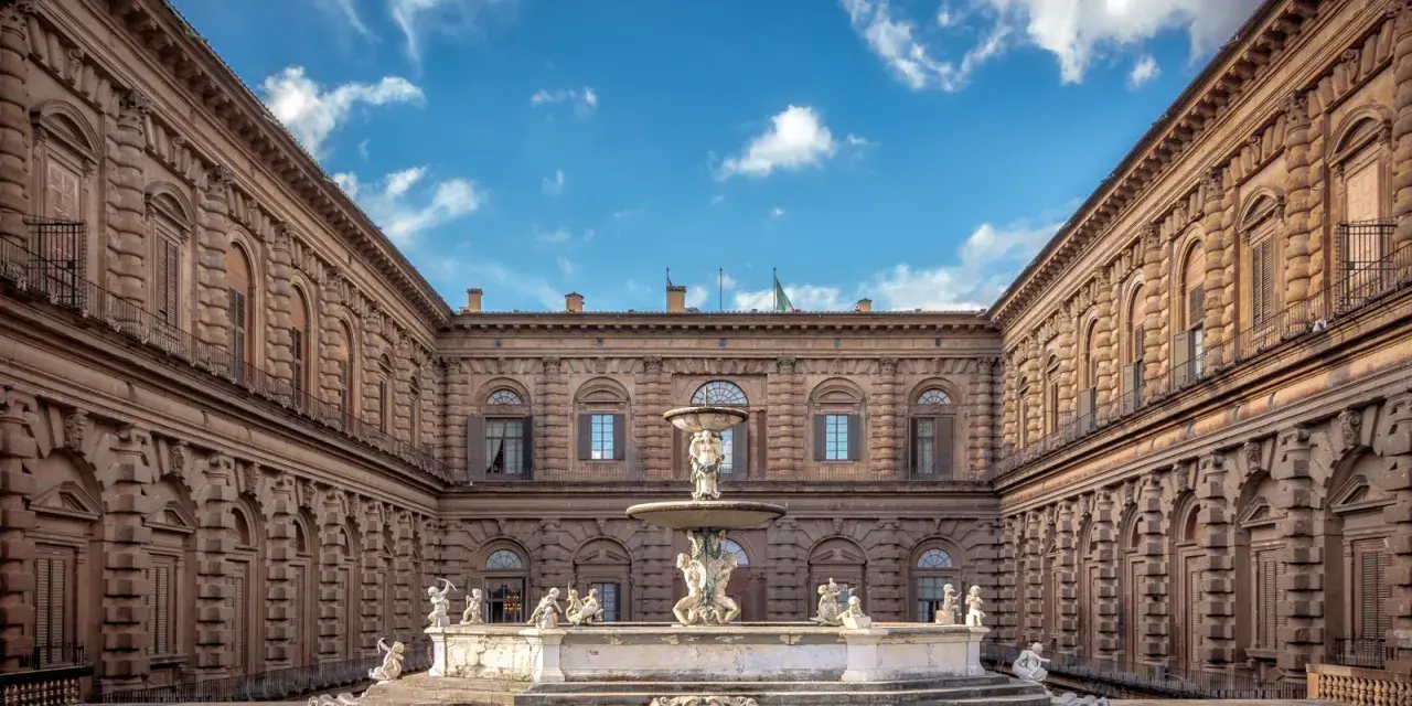 Cortile del Palazzo Pitti con fontana e statue, sotto un cielo azzurro con nuvole bianche.